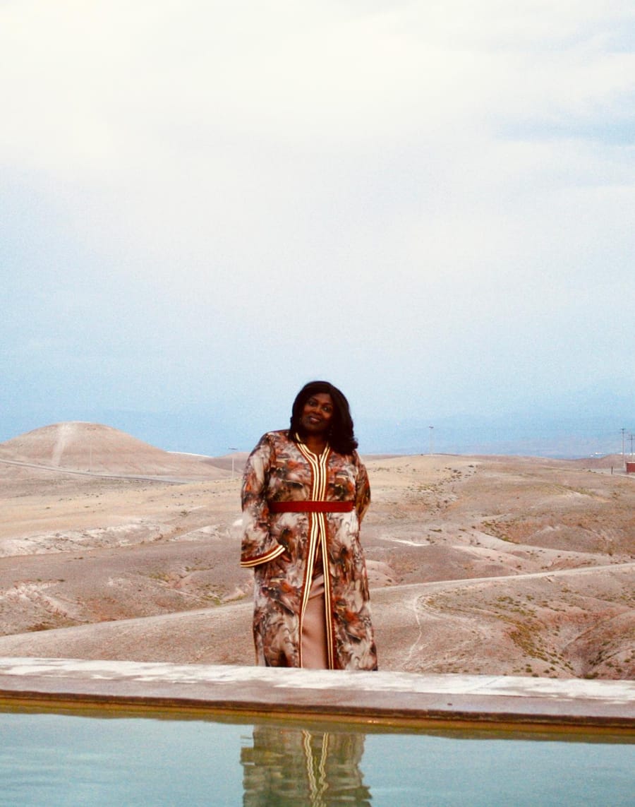 Merley in a patterned kaftan standing by a pool, with desert hills behind her.
