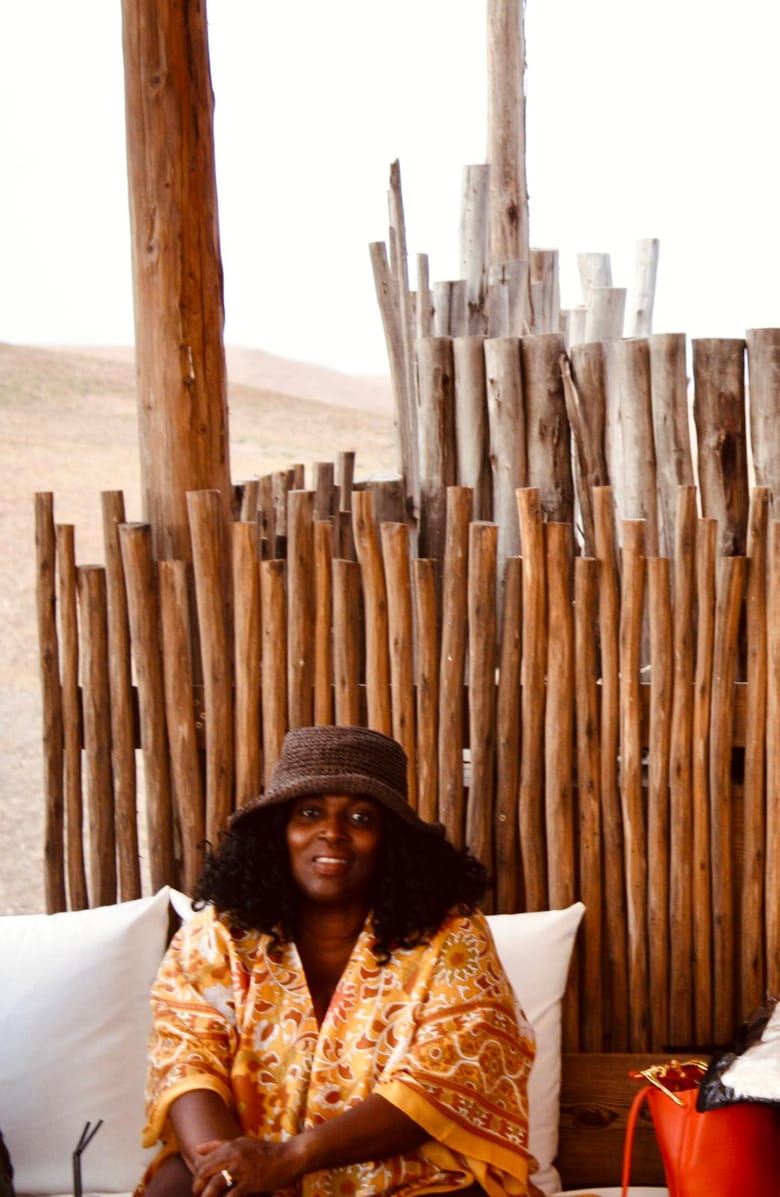 Merley in a wide-brimmed hat and a patterned shirt, seated on white cushions in front of a wooden fence, desert hills behind.