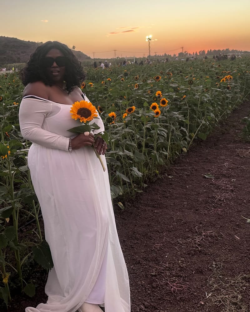 Merley in a white dress among tall sunflowers at dusk, holding a sunflower against her chest.